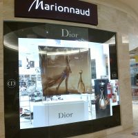 Značení výlohy luxusní prodejny v obchodným centru, laminovaná černá folie s logem Dior lemuje sklo, svítící bílé logo Marionnaud na černé desce nad výlohou, v pozadí další obchody