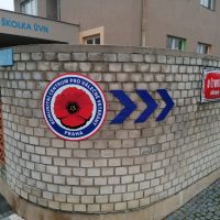 Modro-bílo-červené kulaté logo Komunitního centra pro válečné veterány Praha přilepené na zaoblený roh cihlové zdi v exteriéru, vpravo tři modré navigační šipky
